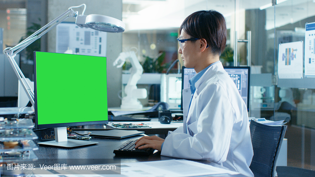 坐在办公桌前的亚洲科学家在一台模拟绿色屏幕的个人电脑上工作。在计算机科学研究实验室的机械臂模型。